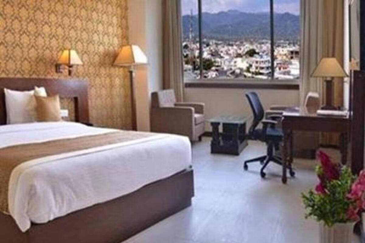 A hotel room with a large bed, a desk, a chair, and a view of a cityscape through a large window.