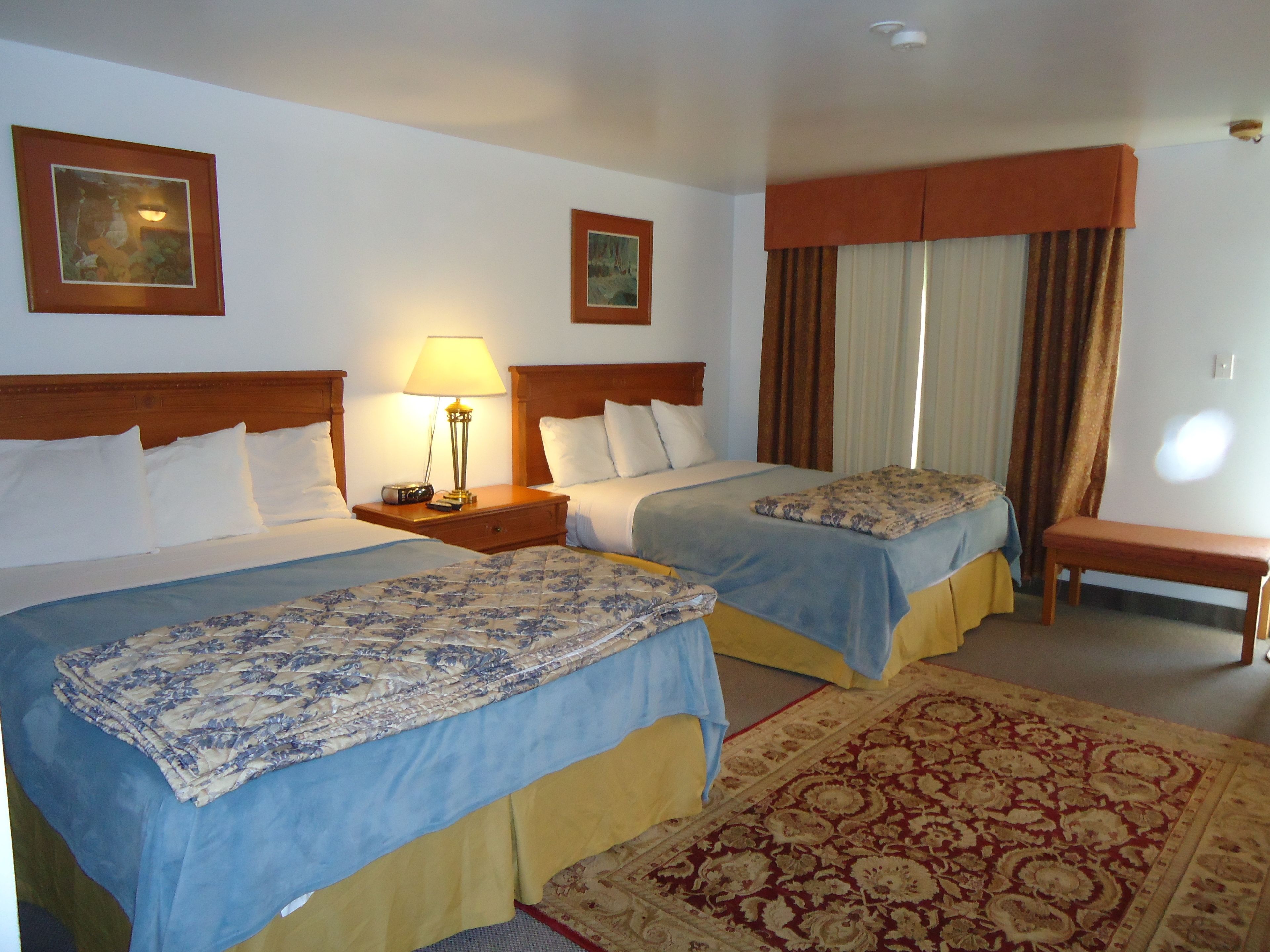 Quarto de hotel com duas camas, uma mesa de cabeceira com abajur, um banco e um tapete grande com padrão.