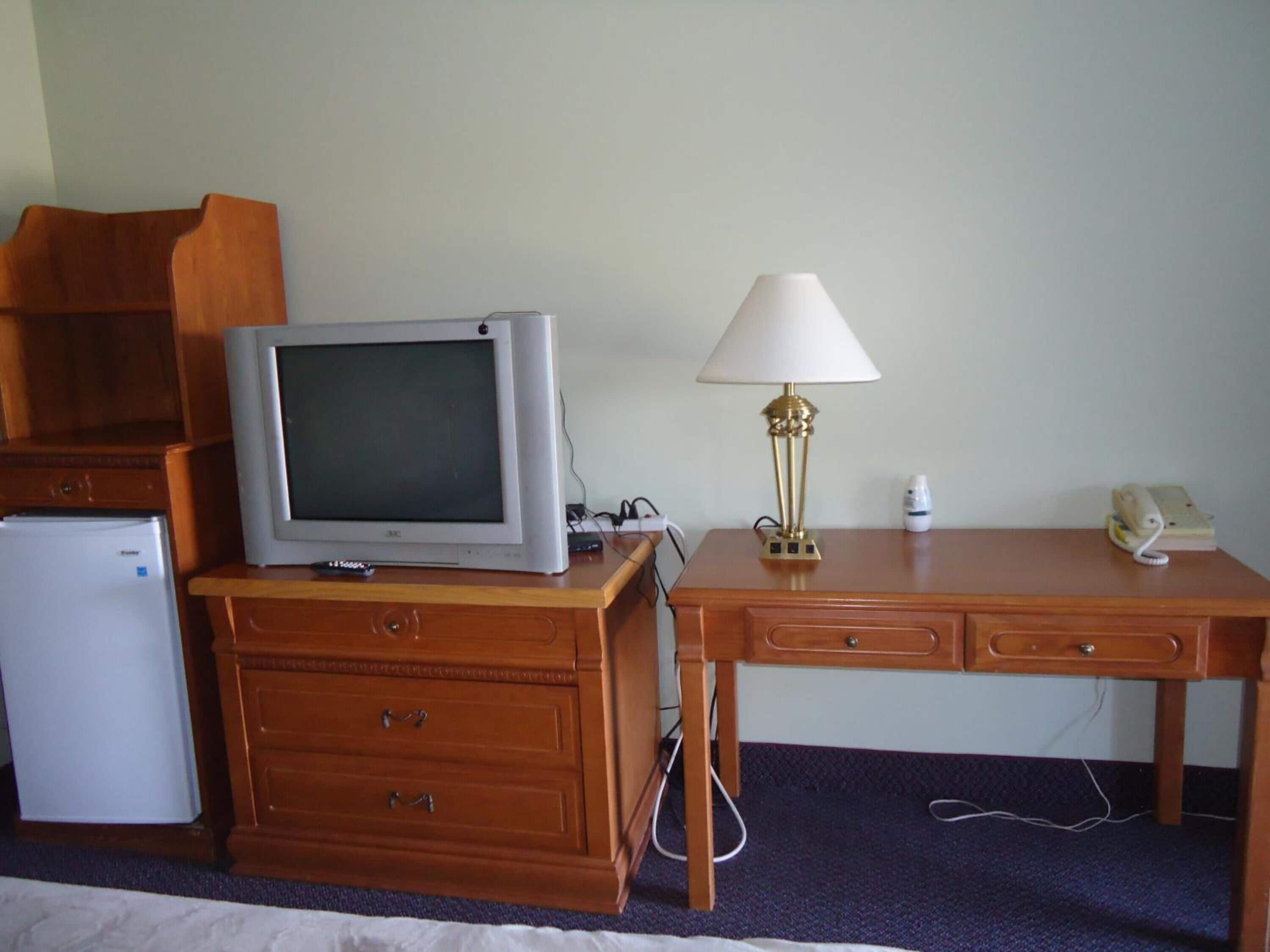 Habitación de hotel con escritorio de madera, televisor, lámpara, teléfono y refrigerador.