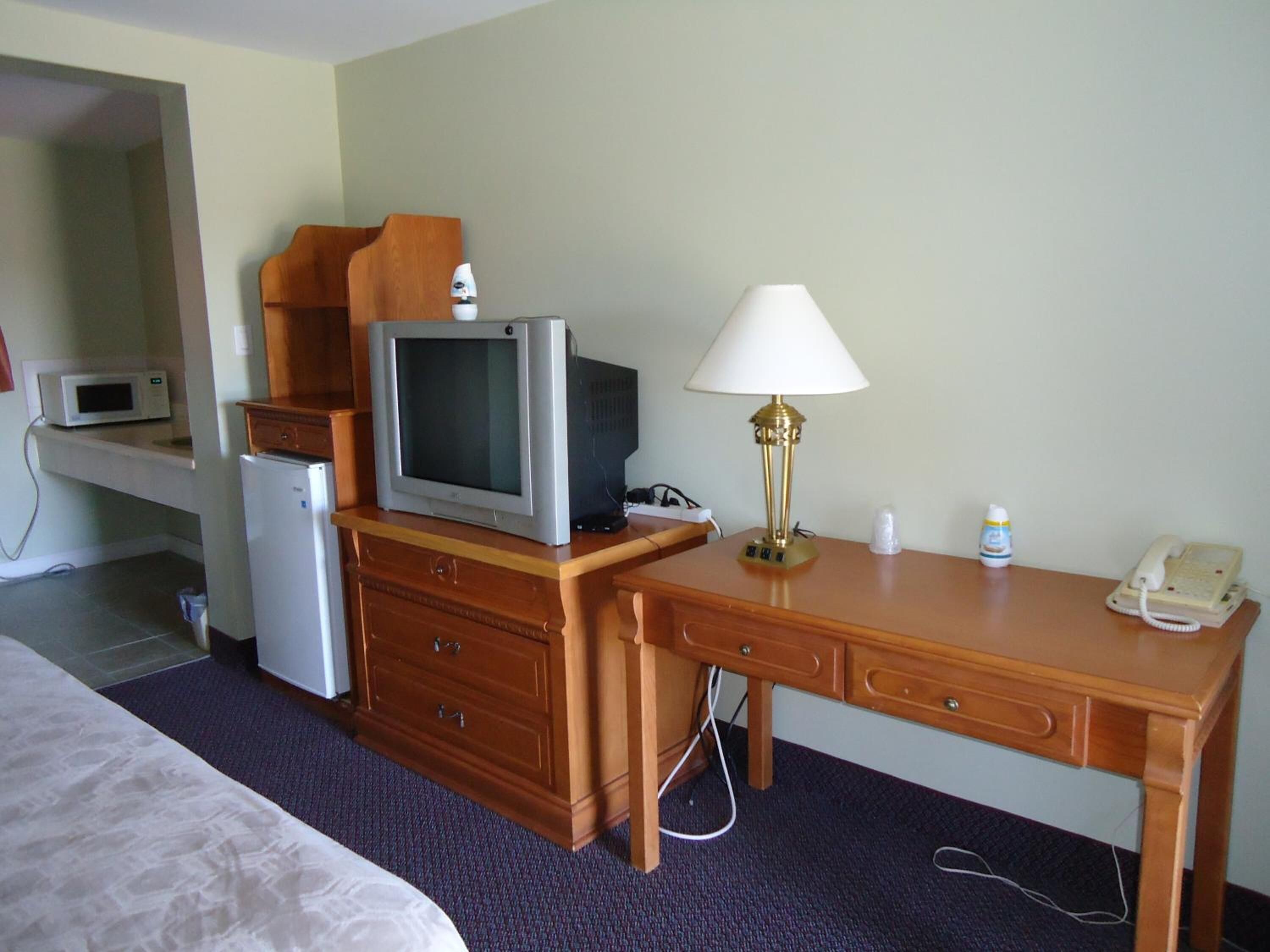 Una habitación de hotel con cama, escritorio de madera, televisor, lámpara, microondas, refrigerador y teléfono.