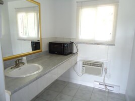 Un baño con lavamanos, espejo, microondas negro y ventana con persianas.