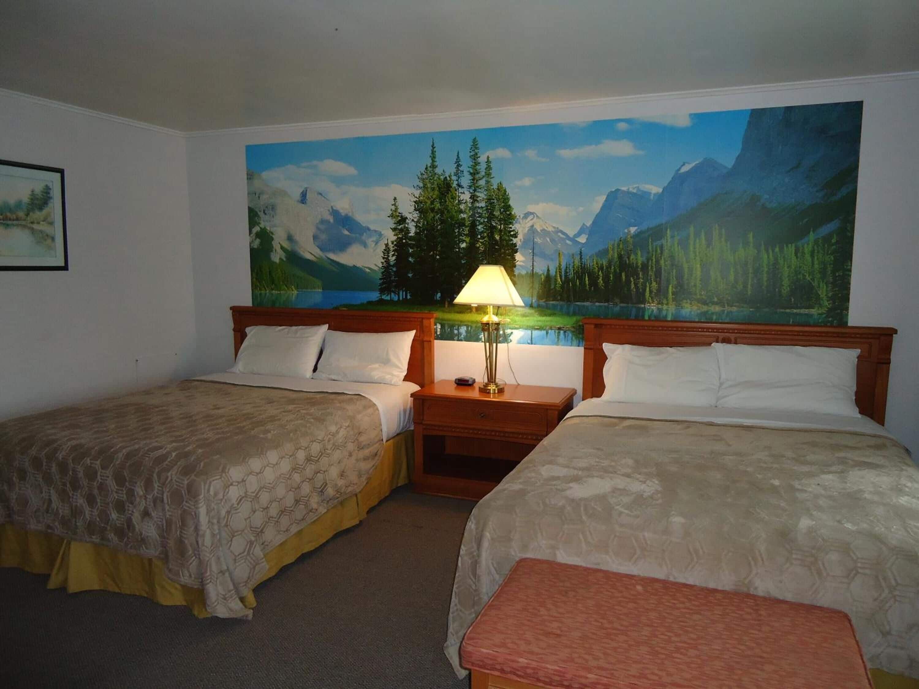Een hotelkamer met twee bedden, een nachtkastje met een lamp, en een groot schilderij van een berglandschap aan de muur.