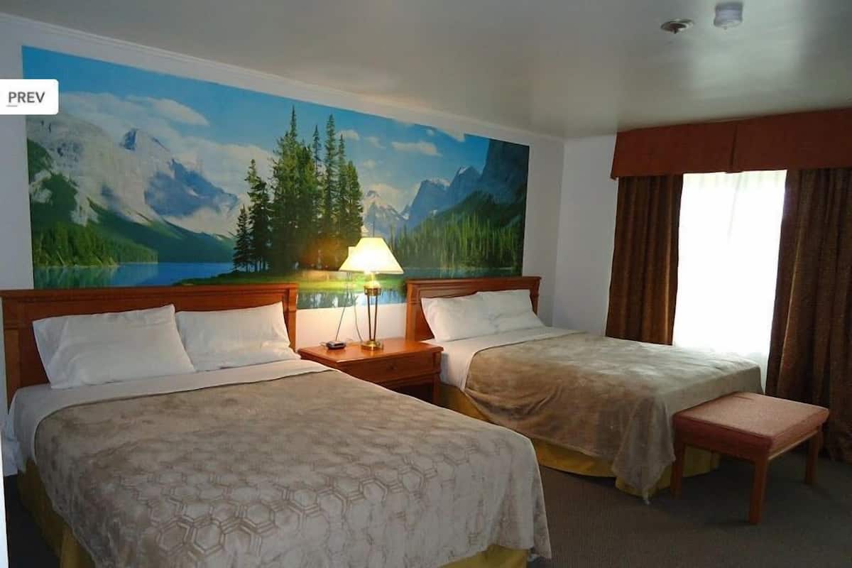 A hotel room with two beds, a nightstand with a lamp, a painting of a mountain landscape, and a window with curtains.