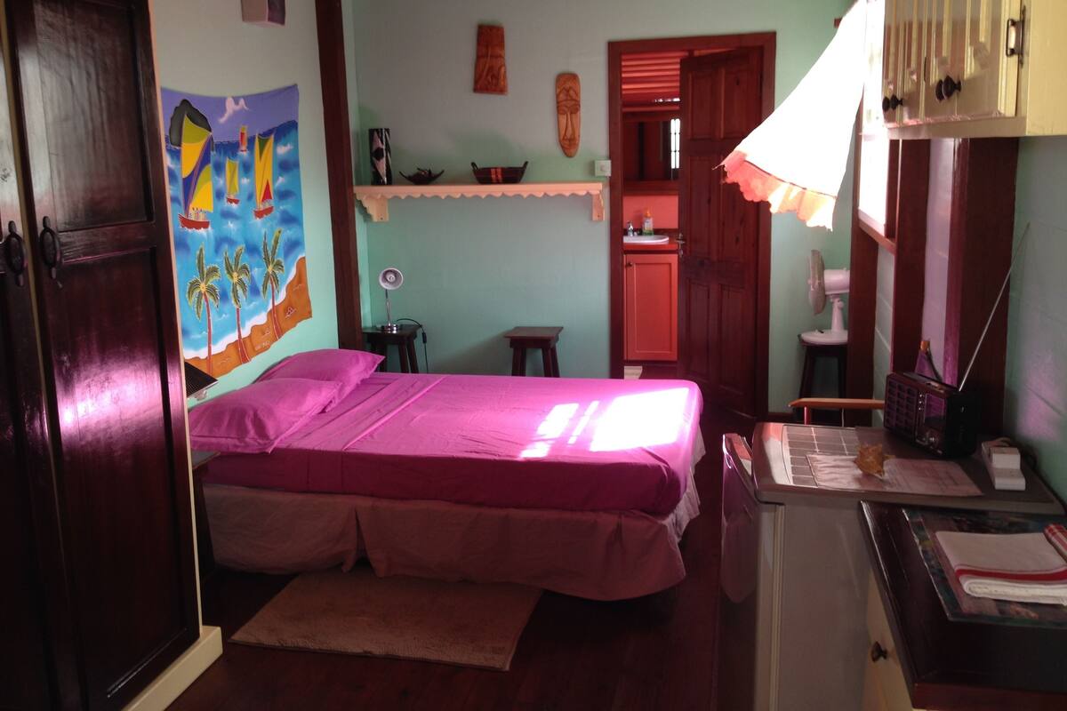 A room with a bed, a desk, a chair, a shelf with items, a door, and a wall with a painting.