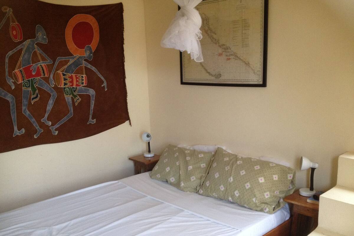 A bed with white linens, a green patterned pillow, a wall tapestry, a map, and a small table with a lamp.