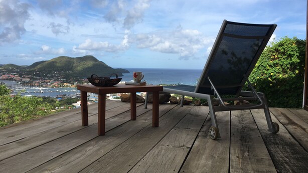 A wooden deck with a table and a chair overlooking a marina and distant hills. - Italian Guesthouse (Gros Islet)