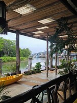 Un espacio al aire libre techado con vista a un río y barcos, pilares de madera y plantas en macetas.