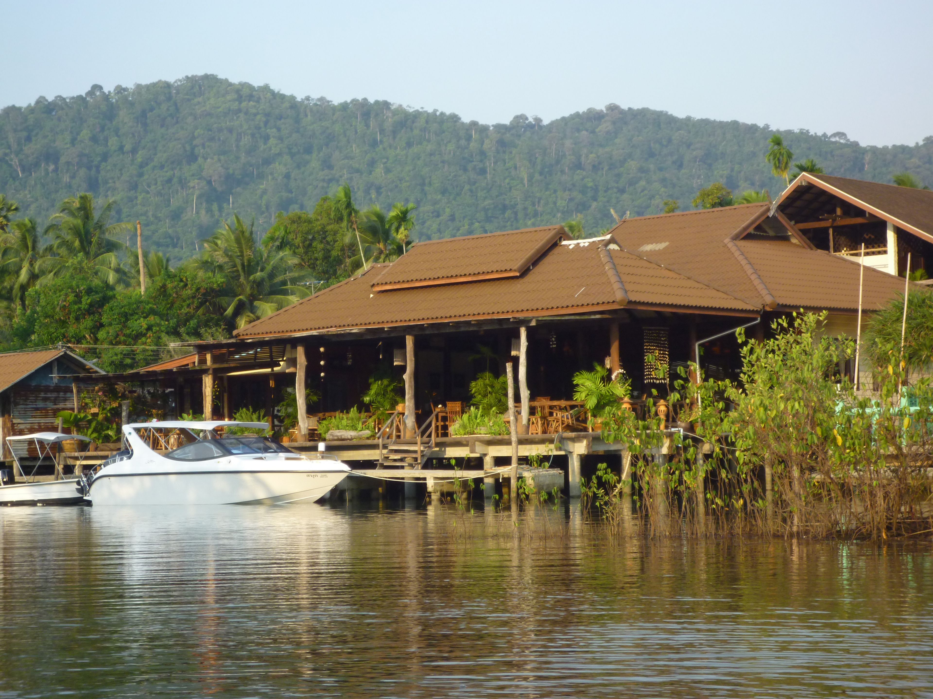 A waterfront property with a boat docked in front, wooden buildings with a rustic design, and a lush green hill in the background.