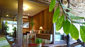 Ein modernes Wohnzimmer mit Holzboden, einem runden Tisch mit Pflanze, einer Couch mit gemusterten Kissen und Blick in die Küche durch eine Glastür.