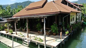 Ein Hausboot mit überdachter Terrasse, Holzmöbeln und Blumentöpfen, liegt in einem ruhigen Gewässer mit Bergen im Hintergrund.