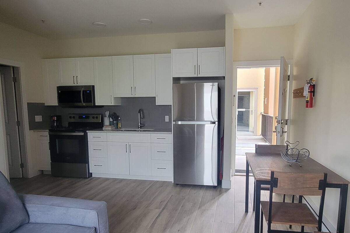 A compact kitchen with white cabinets, a microwave, a stainless steel refrigerator, and a dining area with a wooden table and chairs.