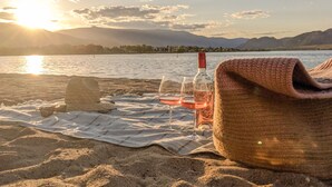 A beach scene with a blanket, a bottle, and glasses on the sand, overlooking a calm lake with mountains in the background. - Desert Surfside Waterfront Accommodations (Osoyoos)