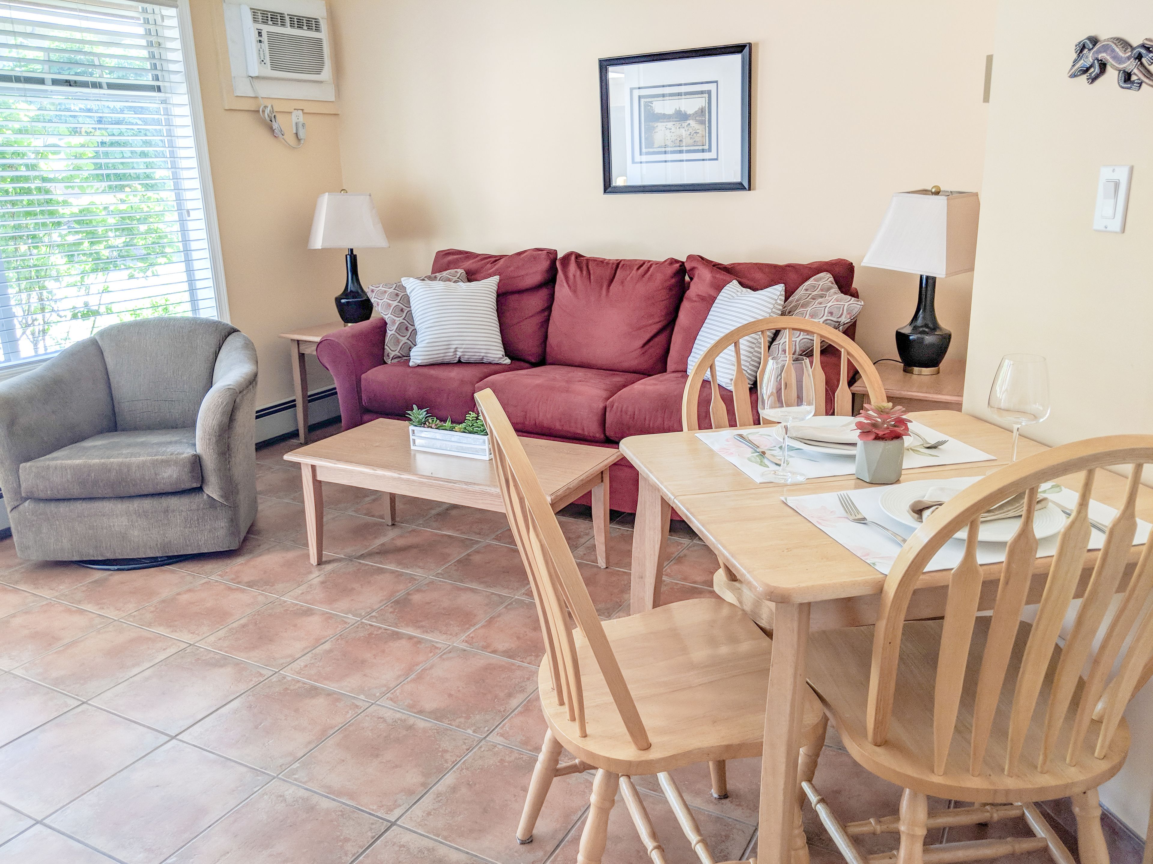 A living room with a sofa, armchair, dining table, chairs, and a small side table with a lamp.