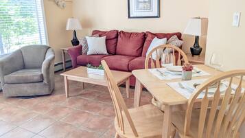 A living room with a sofa, armchair, dining table, chairs, and a small side table with a lamp.
