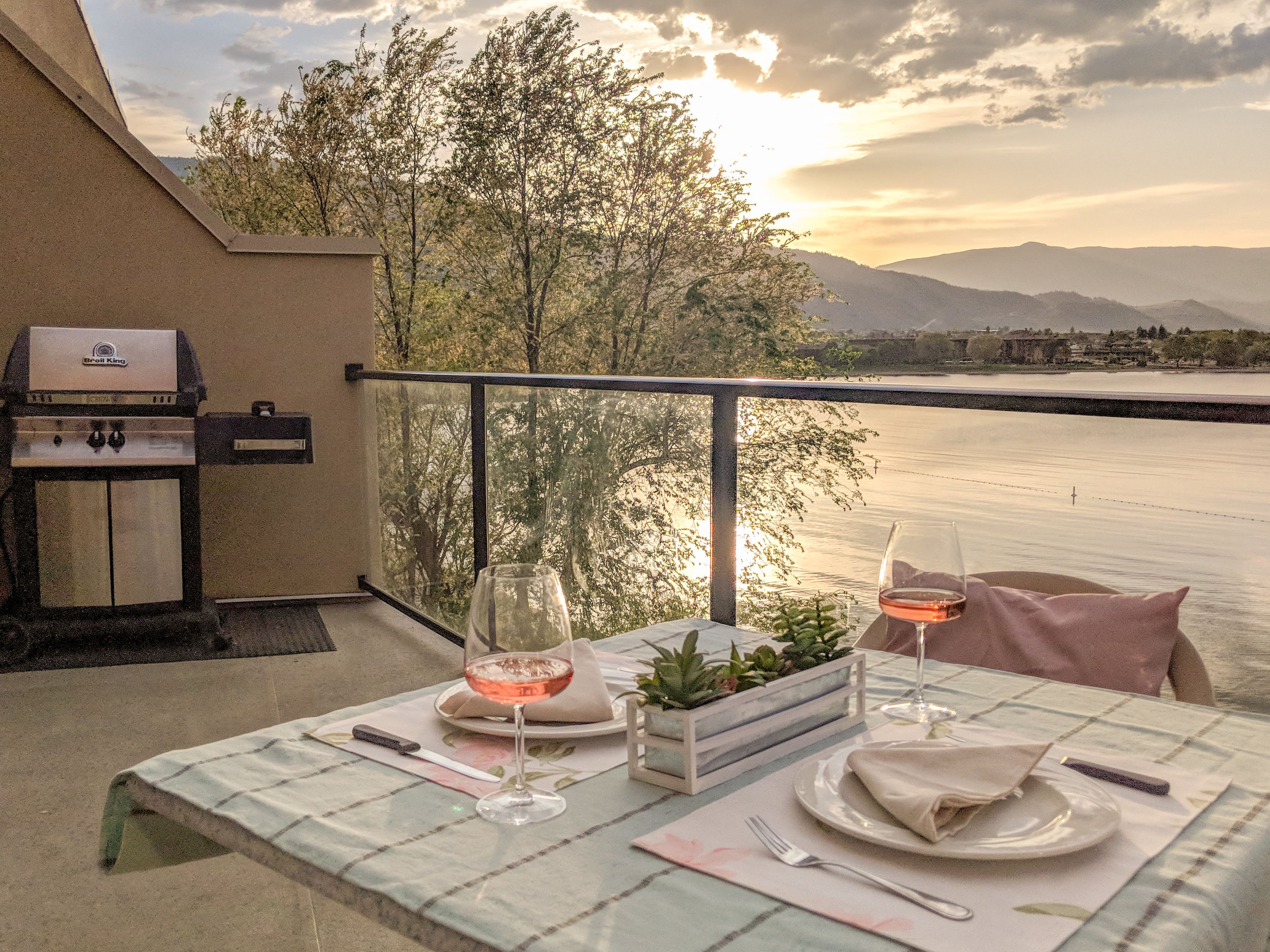 A balcony with a grill, a table set for two with wine glasses, a plant, and a view of a body of water and mountains.