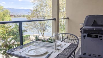 A balcony with a table set for two, a grill, and a view of a lake and mountains.