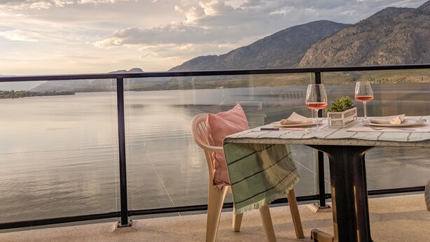 A table with two wine glasses and a small plant, overlooking a calm lake with mountains in the background. - Desert Surfside Waterfront Accommodations (Osoyoos)