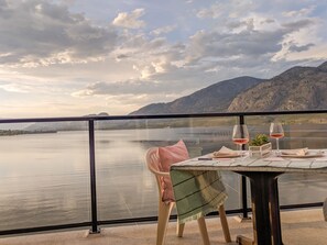 A table with two wine glasses and a small plant, overlooking a calm lake with mountains in the background. - Desert Surfside Waterfront Accommodations (Osoyoos)