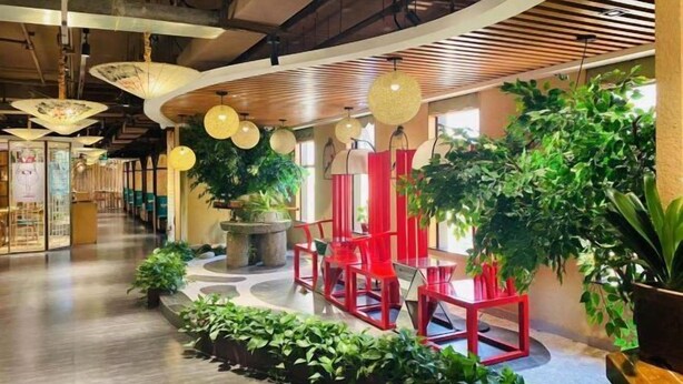 Um interior moderno com uma área de estar central, com cadeiras vermelhas, rodeada por plantas verdes exuberantes e luminárias decorativas pendentes.