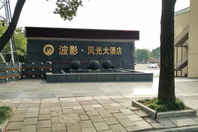 A building with a sign that reads '波影·风光大酒店' and a paved area with a bench and a tree.