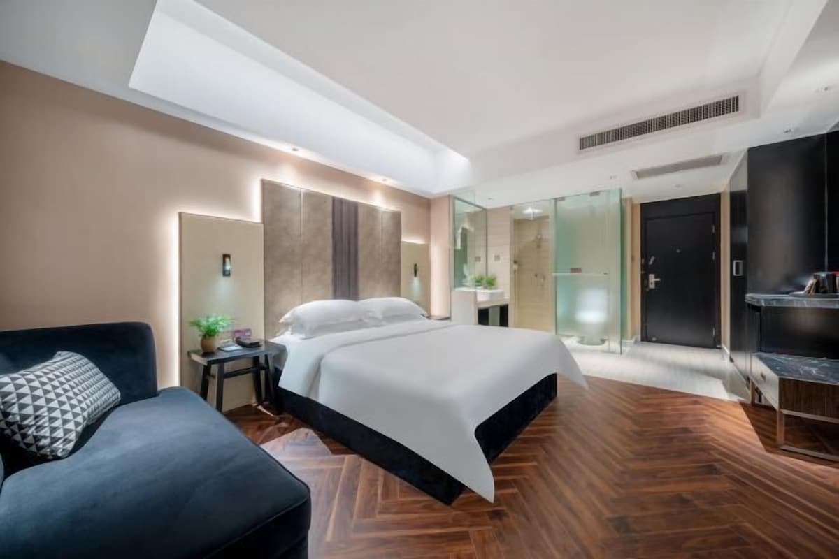 Une chambre d’hôtel moderne avec un grand lit, un canapé, une petite table avec une plante et une douche en verre.