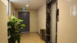 A hallway with a marble floor, a potted plant, a door with an emergency exit sign, and a wall-mounted elevator control panel. - Greentree Inn Shanghai Baoshan Yanghang Shuichan Road Hotel (Shanghai)