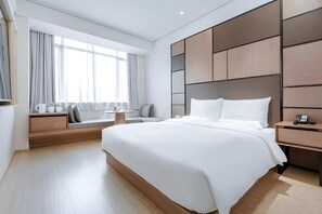 A modern hotel room with a large bed, a bedside table with a phone, a nightstand, a seating area with a bench and cushions, and a window with curtains. - Ji Hotel (Shanghai)