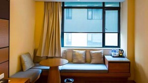 Ein gemütliches Hotelzimmer mit einer Sitzecke, einem kleinen runden Tisch, einem Nachttisch mit Lampe und einem Fenster mit Vorhängen.