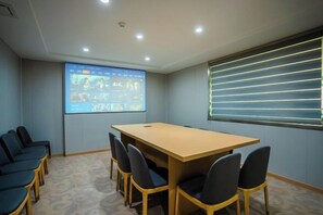 Un salón de conferencias con una larga mesa, sillas, una pantalla para proyector y un ventanal con persianas.