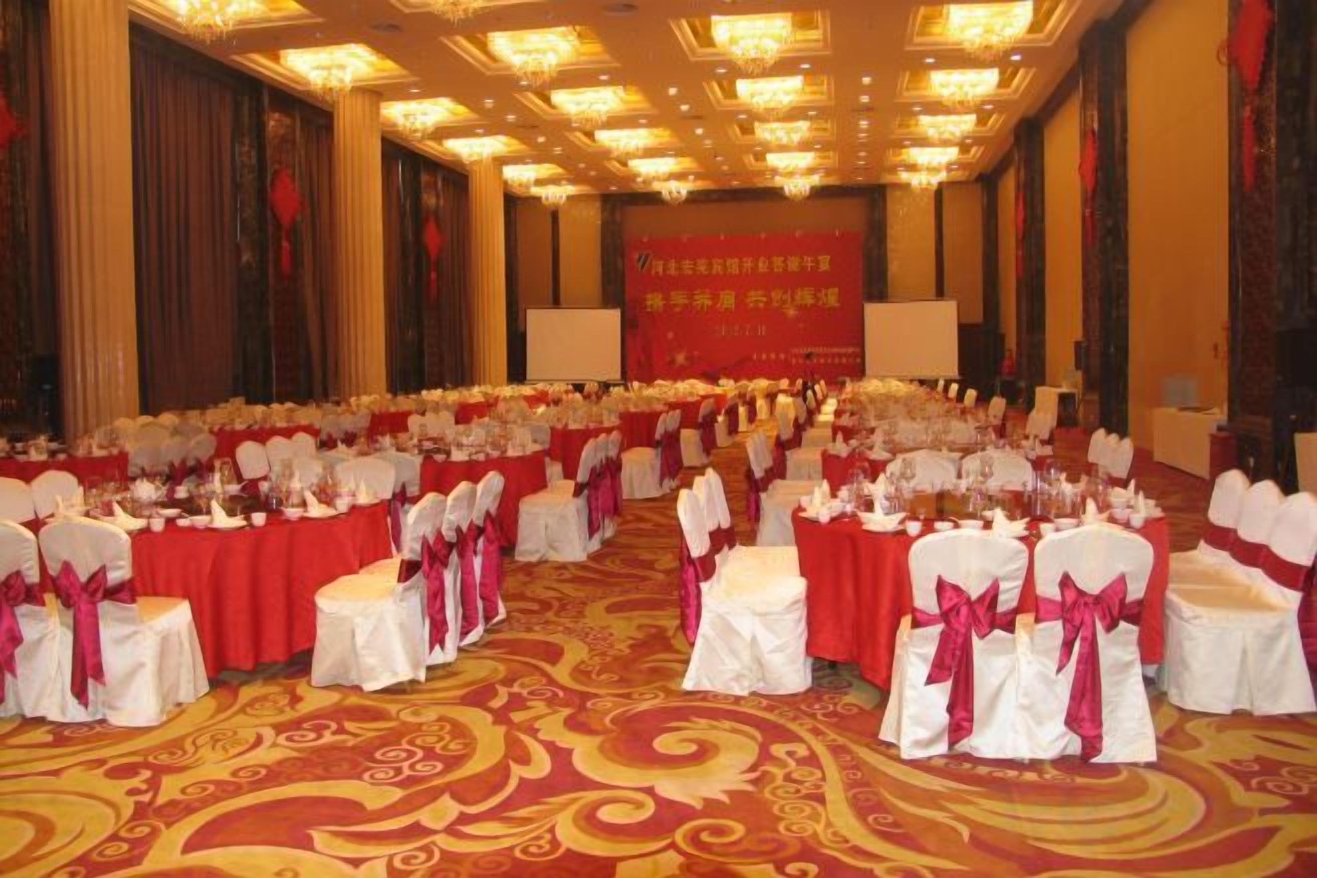 Un salón de banquetes con mesas redondas, sillas blancas y manteles rojos.