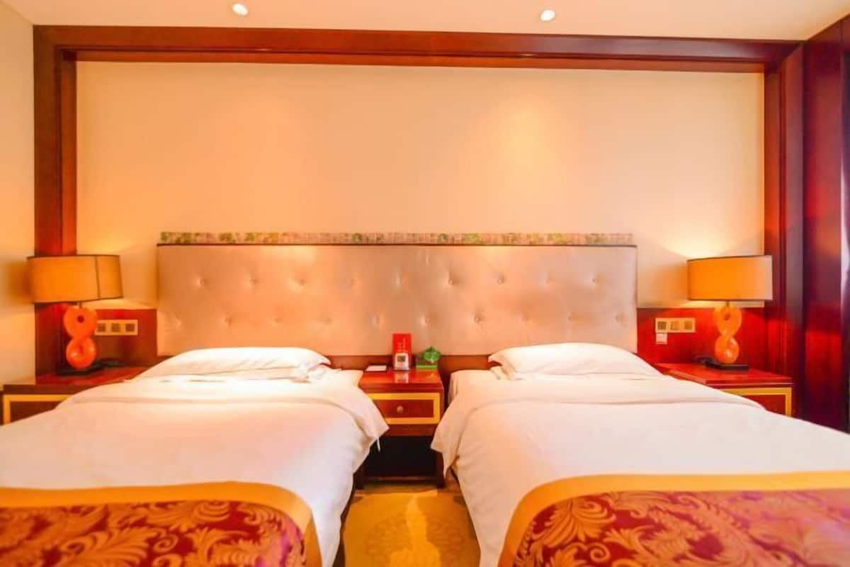 A hotel room with two beds, a large padded headboard, bedside lamps, and a small table with a red lamp.