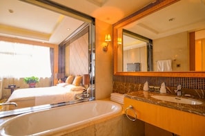 Habitación de hotel con una bañera grande, una cama, un baño con lavamanos y espejo, y vistas al exterior.