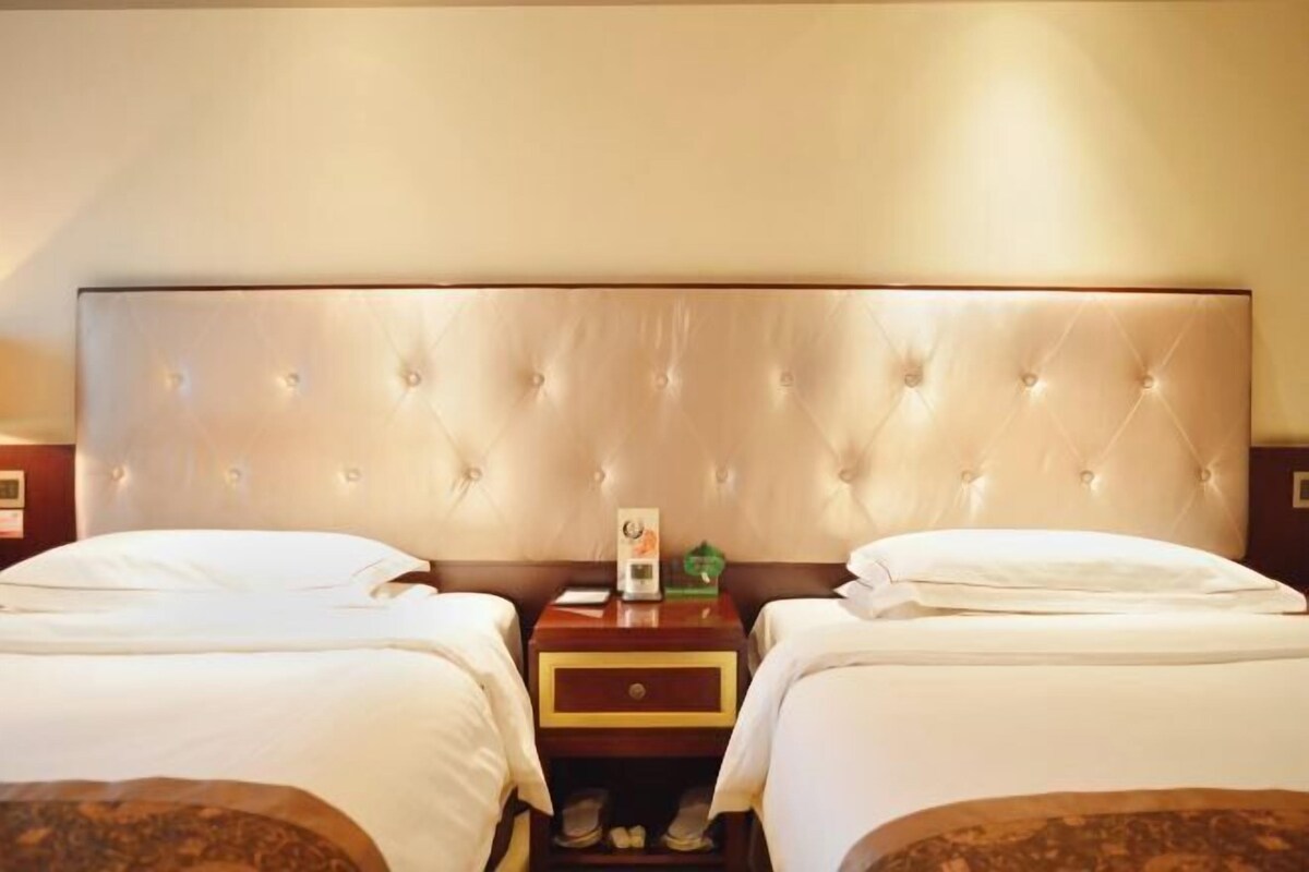 A hotel room with two beds, a large tufted headboard, a nightstand with a clock and a green object, and a wooden drawer.