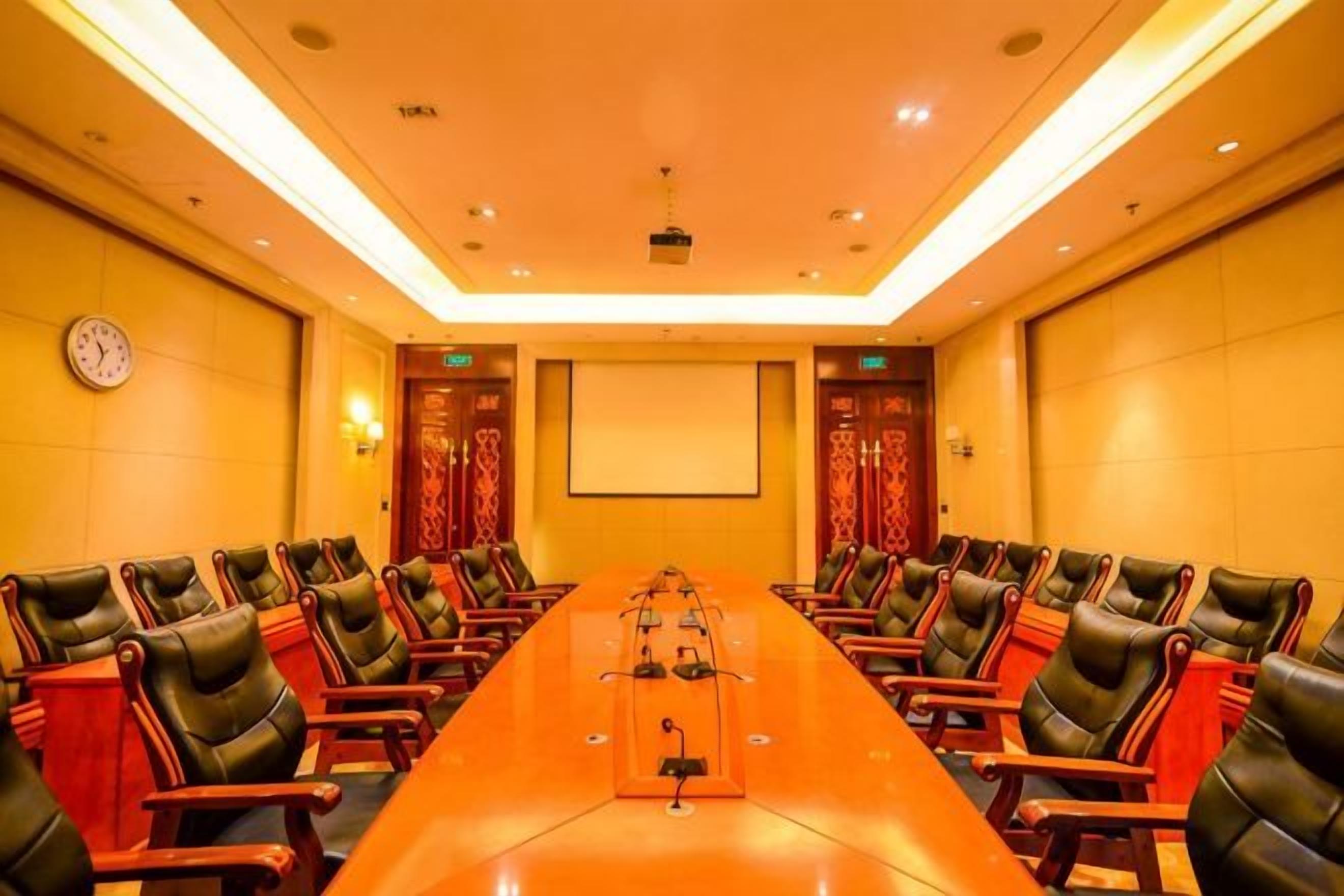 A conference room with a long table, multiple chairs, a projector screen, and a clock on the wall.