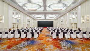 Un amplio salón de conferencias con filas de mesas y sillas, una pantalla grande y candelabros decorativos.