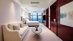 Un cuarto de hotel con una cama grande, un sofá, una mesa de centro redonda con fruta, un cuadro en la pared y vista a la ciudad por la ventana.