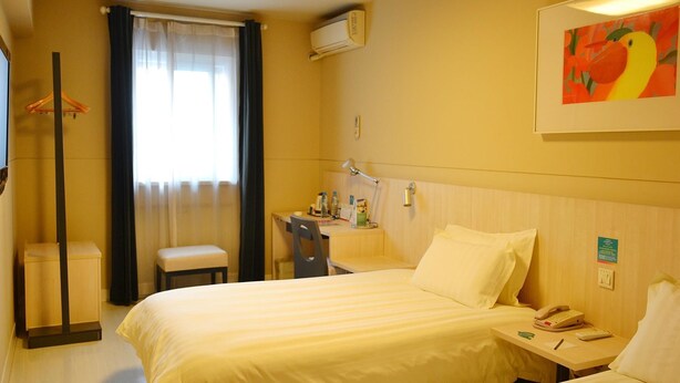 Ein Hotelzimmer mit zwei Betten, einem Schreibtisch mit Telefon und Lampe, einem Sessel, einem Kleiderhaken und einem Fenster mit Vorhängen.