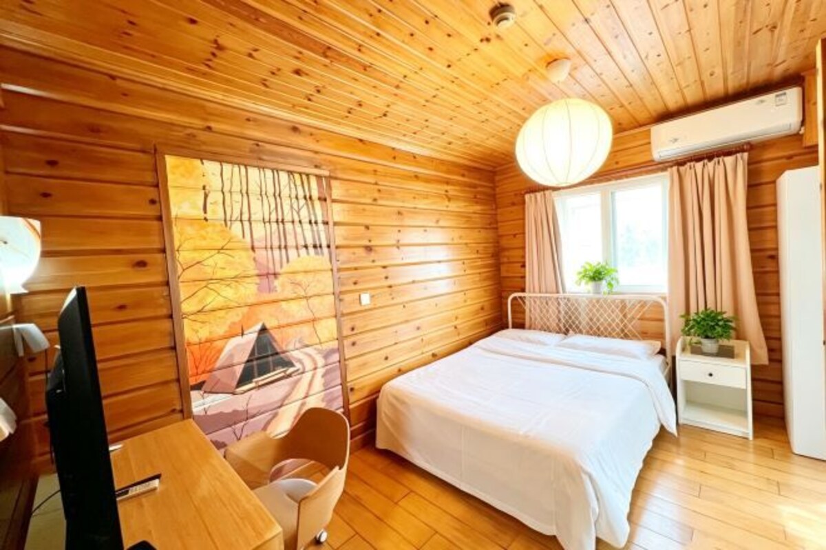 Ein Zimmer einer Holzhütte mit einem Bett, einem Schreibtisch, einem Stuhl, einem Fernseher und einem Fenster mit Vorhängen.