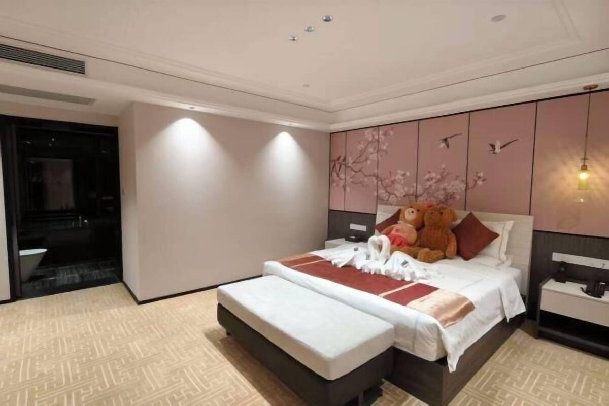 Une chambre d’hôtel avec un lit, une table de chevet, une commode, un grand ours en peluche et un mur décoré d’œuvres d’art représentant des fleurs de cerisier.