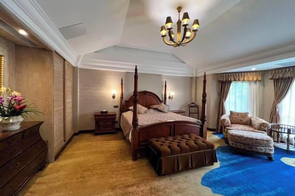 Une chambre à coucher avec un lit à baldaquin, un coin salon avec un fauteuil, une commode et une grande fenêtre avec des rideaux.