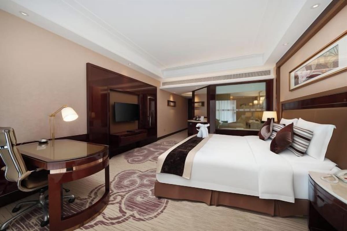A hotel room with a bed, a desk, a chair, a TV, and a bathroom visible through an open door.