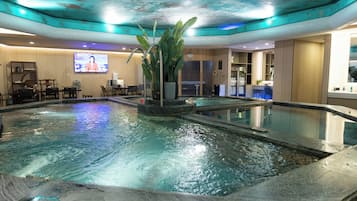 A hotel lobby with a large indoor swimming pool, a television screen, and a reception desk.