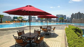 A poolside area with tables and chairs, umbrellas, and a cityscape in the background.