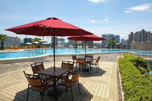 Een zwembadgedeelte met tafels, stoelen, parasols en een stadsgezicht op de achtergrond.