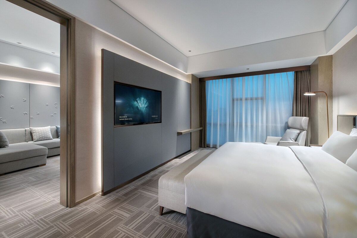 Une chambre d’hôtel moderne avec un grand lit, un canapé, un téléviseur fixé au mur et une vue sur le ciel par la fenêtre.