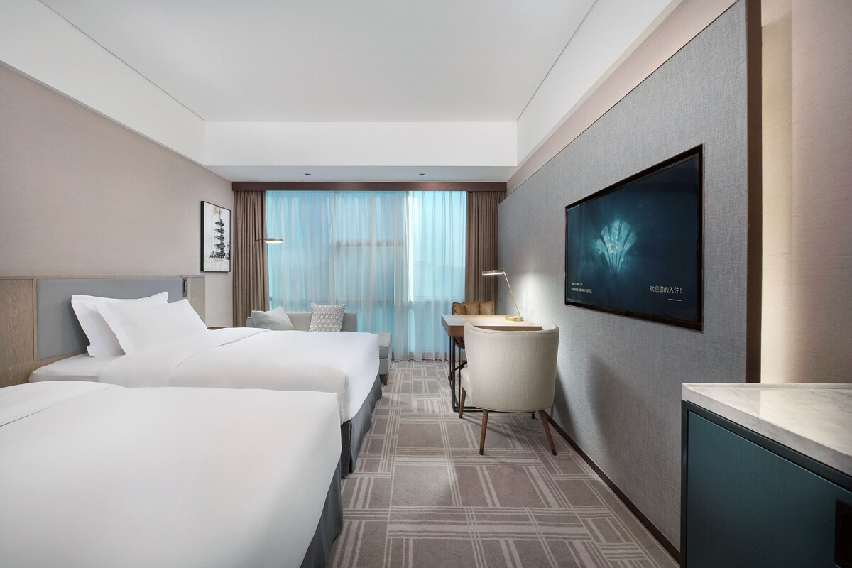 Une chambre d’hôtel avec deux lits, un bureau, une chaise, une télévision et une grande fenêtre avec des rideaux.