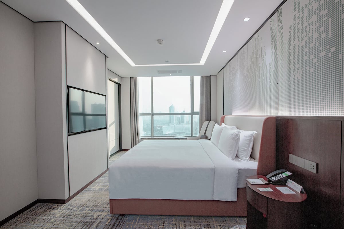 Ein modernes Hotelzimmer mit einem großen Bett, einem flachbildfernseher an der Wand, einem Schreibtisch mit Telefon und Blick auf die Stadt durch große Fenster.