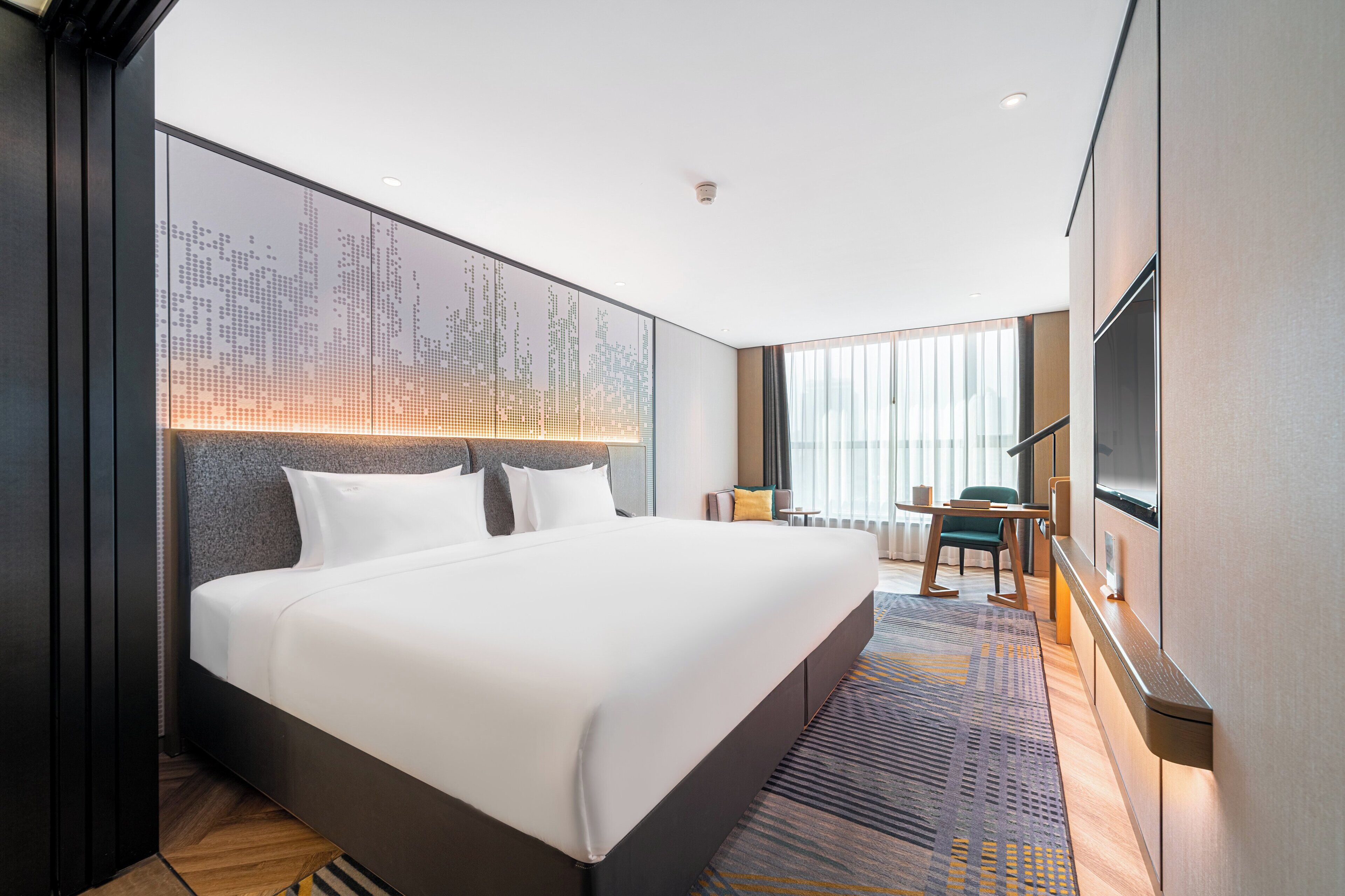 Una habitación de hotel moderna con una cama grande, un cuadro con vista a la ciudad, un escritorio con una silla y un televisor montado en la pared.