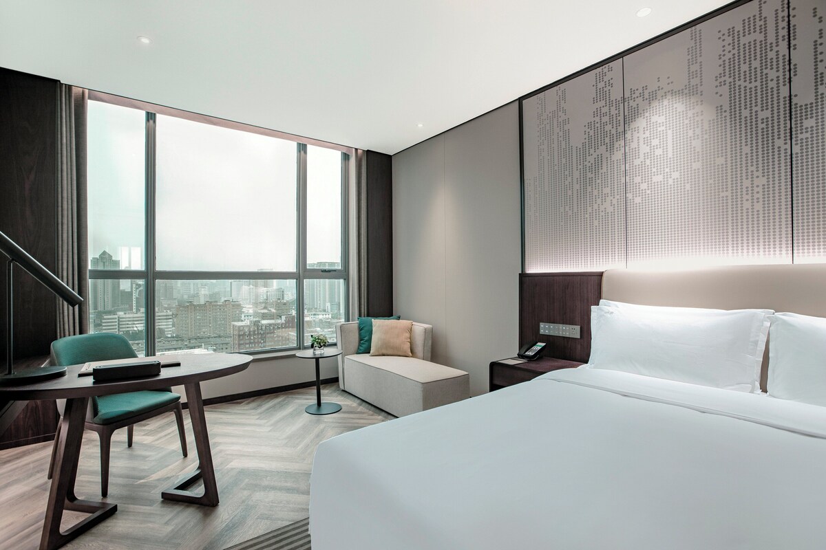 A modern hotel room with a large bed, a seating area with a table and chairs, a small side table, and a large window offering a city view.