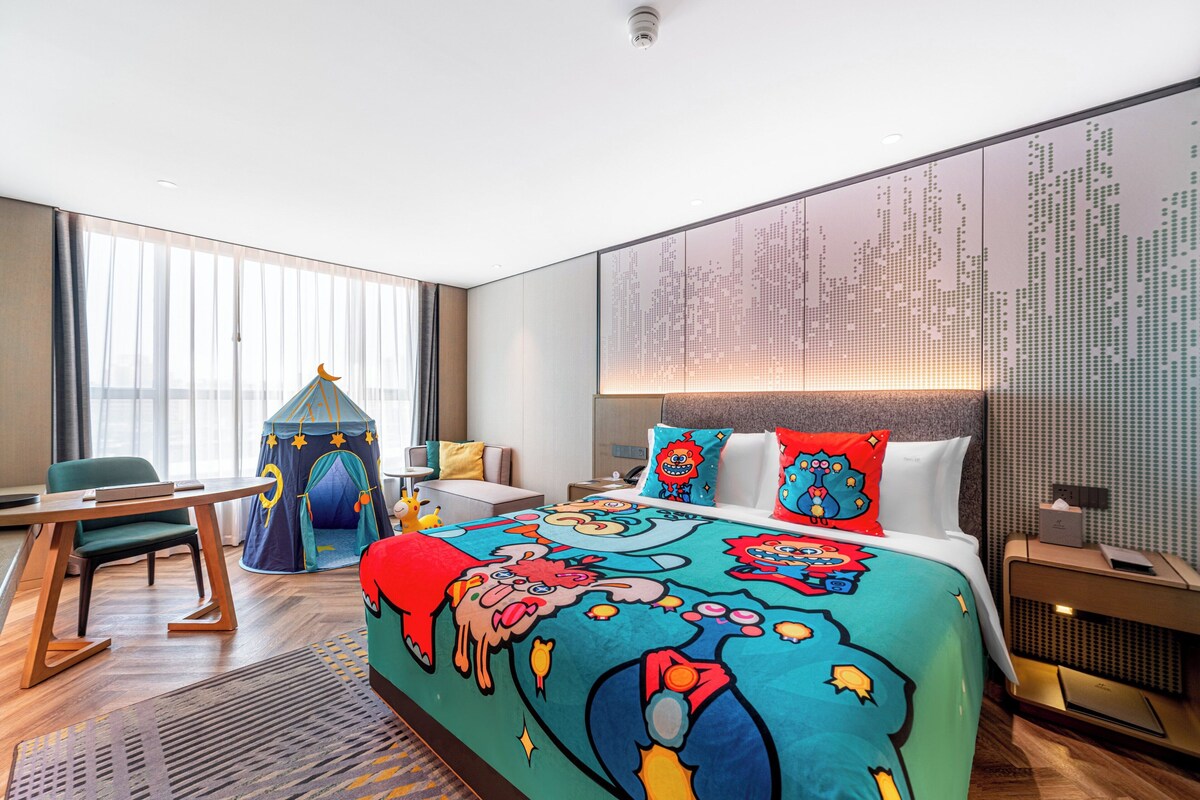 A hotel room with a large bed, a desk, a chair, a play tent, and a wall with a digital display.